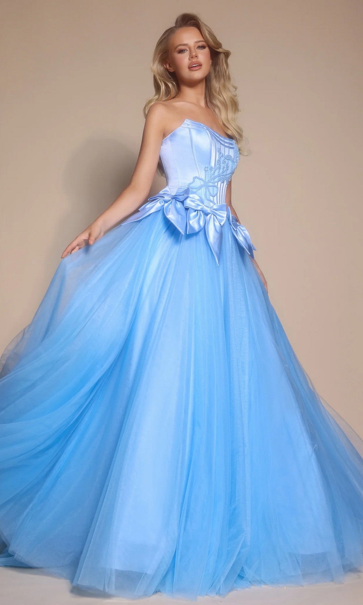 A woman in the light blue Portia and Scarlett PS26102 strapless ball gown, featuring a corset bodice and voluminous tulle skirt, stands gracefully against a plain beige background.